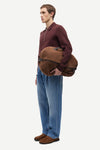 A man in a maroon half-zip pullover, blue jeans, and brown suede shoes is shown in profile holding a brown suede shoulder bag with black trim against a light gray studio background.