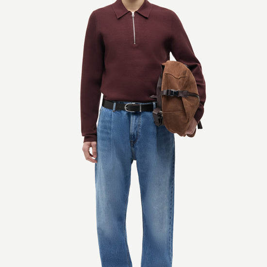 A male model stands against a light gray studio background wearing a burgundy half-zip long-sleeve polo shirt, blue jeans, brown suede loafers, and holding a brown leather hat against his side.
