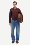 A male model stands against a light gray studio background wearing a burgundy half-zip long-sleeve polo shirt, blue jeans, brown suede loafers, and holding a brown leather hat against his side.