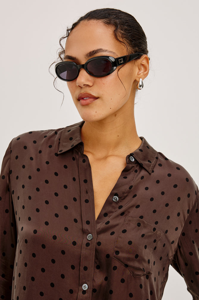 Woman wearing a brown polka dot shirt with sunglasses on a plain background