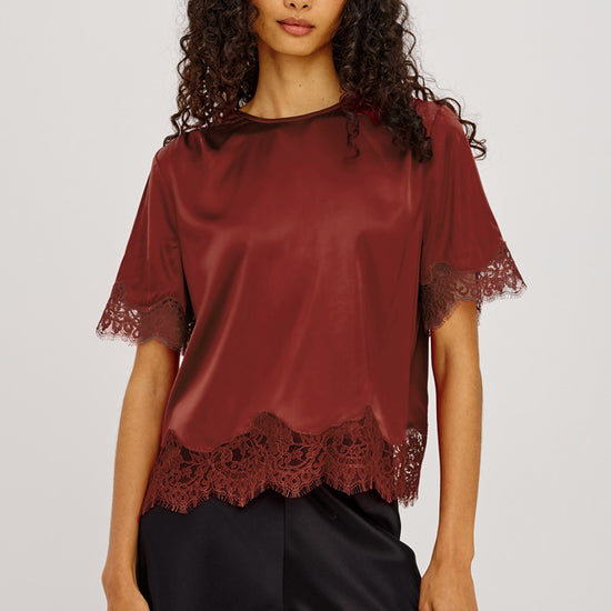 A woman modeling a short-sleeve satin blouse in a deep burgundy color, featuring lace trim at the sleeve cuffs and hem, paired with black satin pants against a light gray studio background.