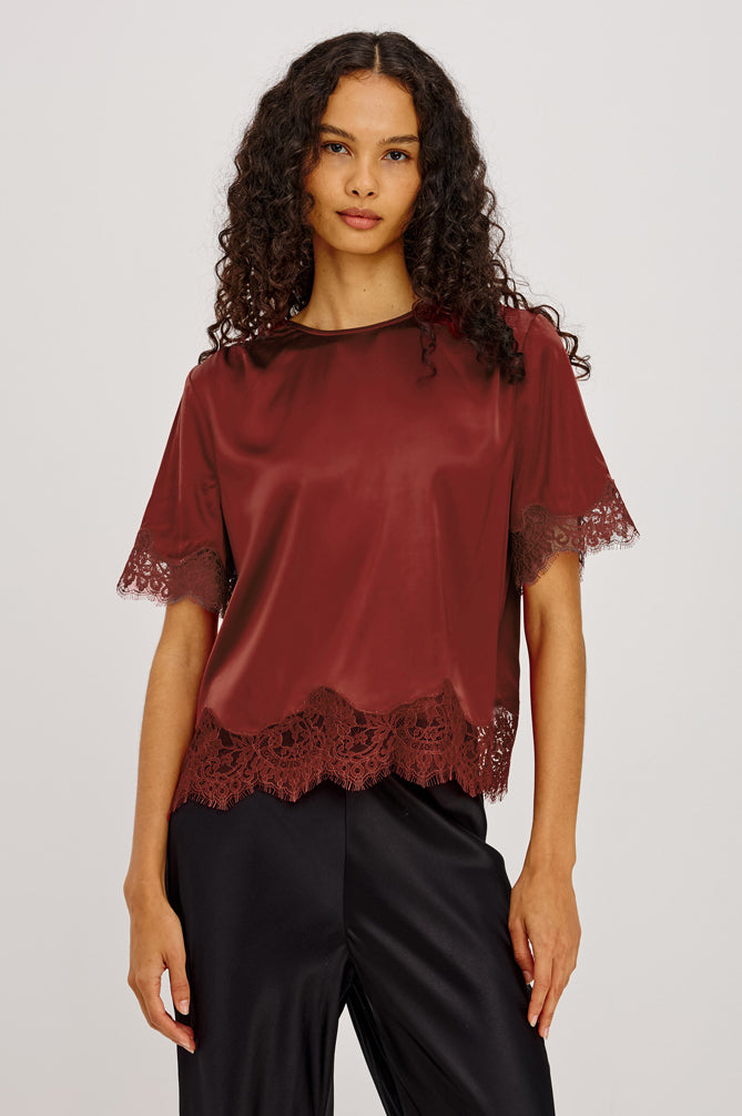 A woman modeling a short-sleeve satin blouse in a deep burgundy color, featuring lace trim at the sleeve cuffs and hem, paired with black satin pants against a light gray studio background.