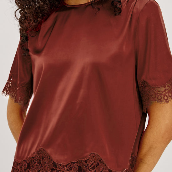 A woman wearing a rust-brown satin short-sleeve top with dark lace trim at the hem and sleeve cuffs, paired with black bottoms, against a light gray studio background.