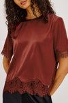A woman wearing a rust-brown satin short-sleeve top with dark lace trim at the hem and sleeve cuffs, paired with black bottoms, against a light gray studio background.
