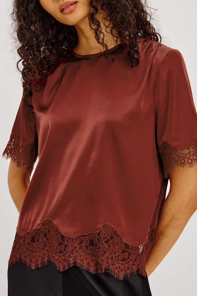 A woman wearing a rust-brown satin short-sleeve top with dark lace trim at the hem and sleeve cuffs, paired with black bottoms, against a light gray studio background.