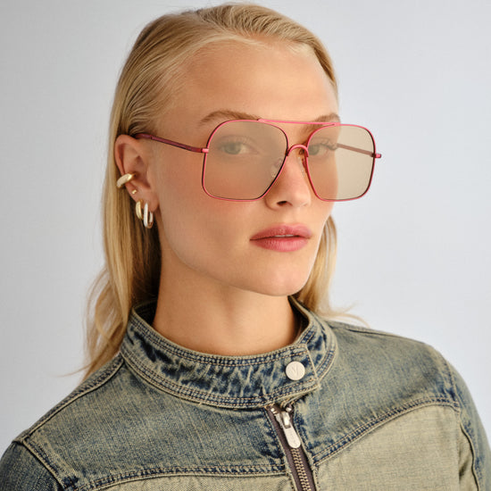 Person wearing pink sunglasses and a denim jacket on a plain background