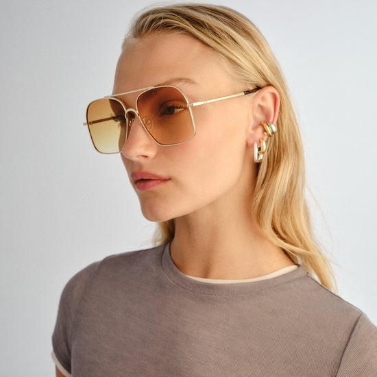 Woman wearing gold sunglasses and earrings on a plain background