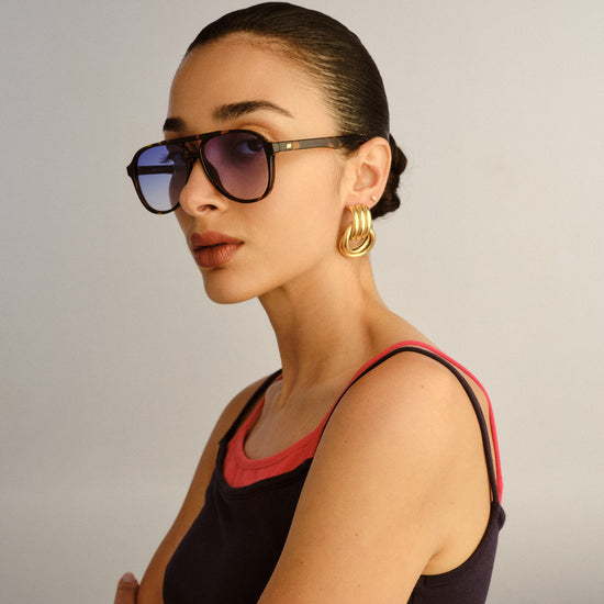 Woman wearing sunglasses and gold hoop earrings against a plain background