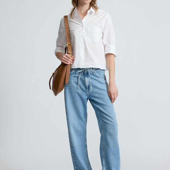 Woman wearing a white shirt and blue jeans holding a brown leather bag on a light gray background