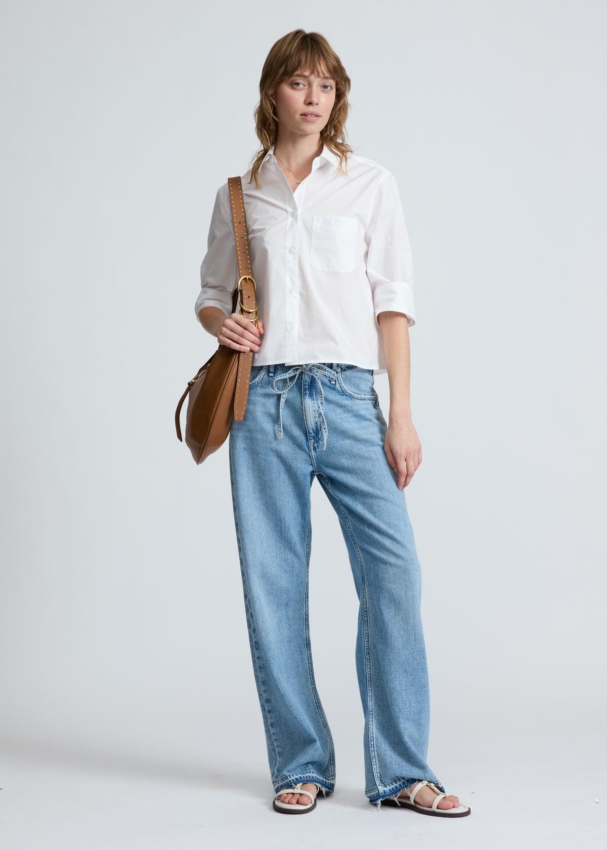 Woman wearing a white shirt and blue jeans holding a brown leather bag on a light gray background