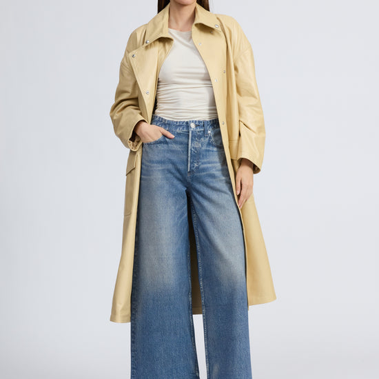 Woman wearing a beige trench coat, white shirt, blue jeans, and black boots on a light gray background