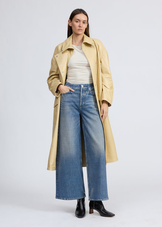 Woman wearing a beige trench coat, white shirt, blue jeans, and black boots on a light gray background