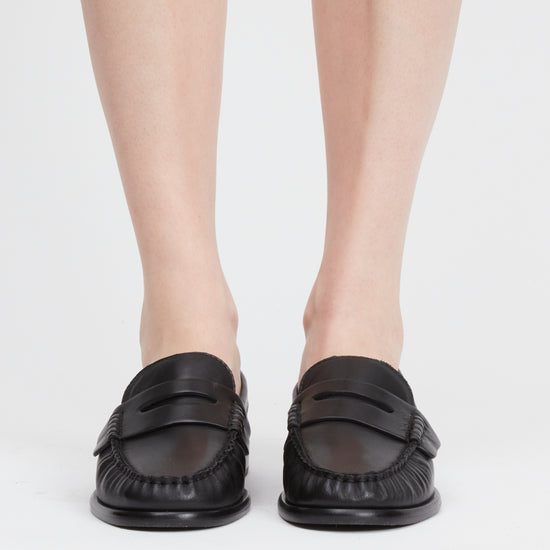A pair of black leather penny loafers worn by a model, shown from the front against a clean white studio background.