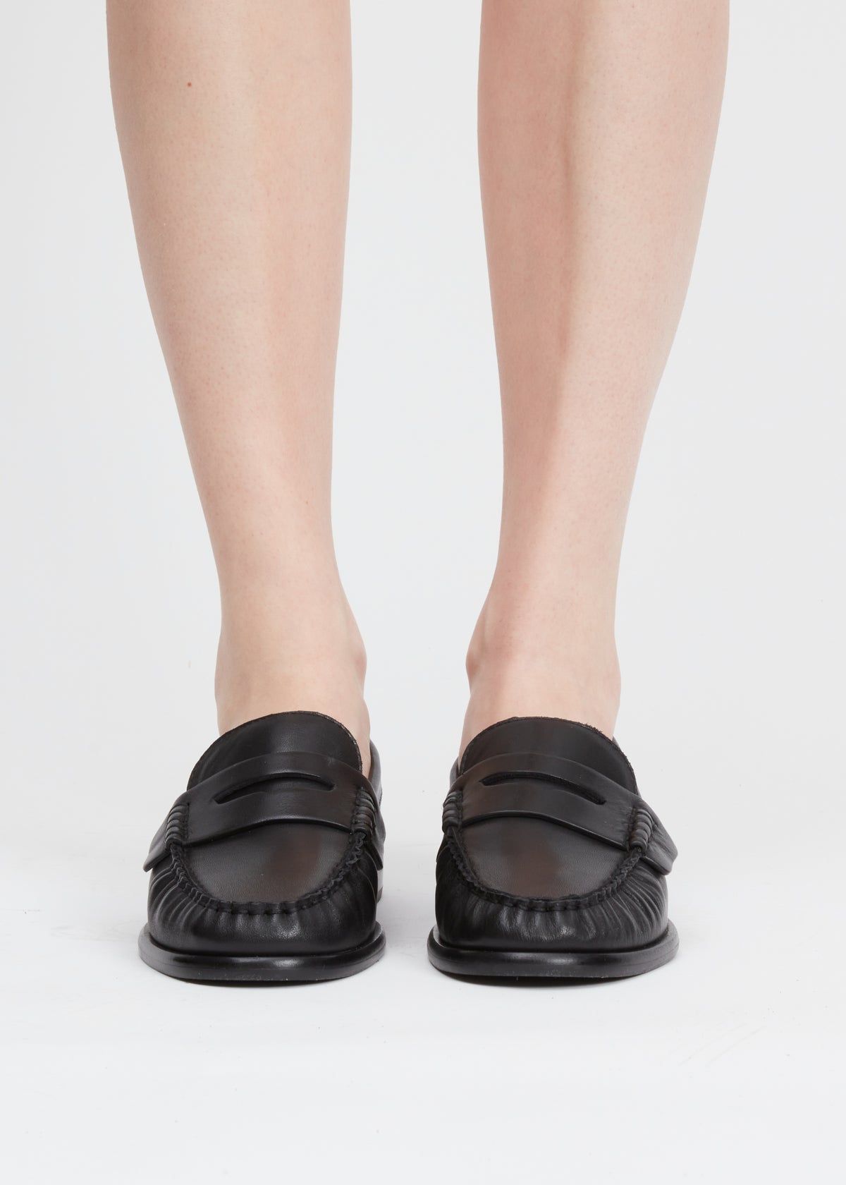 A pair of black leather penny loafers worn by a model, shown from the front against a clean white studio background.