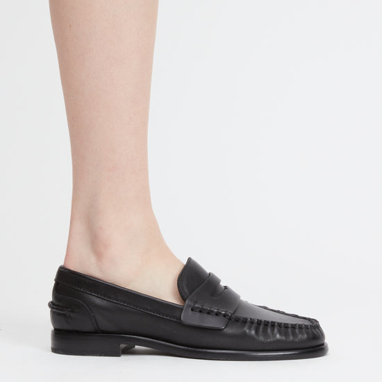 A person's foot wearing a black leather penny loafer with fringe detailing on the toe, shown against a plain light gray background.