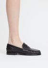 A person's foot wearing a black leather penny loafer with fringe detailing on the toe, shown against a plain light gray background.