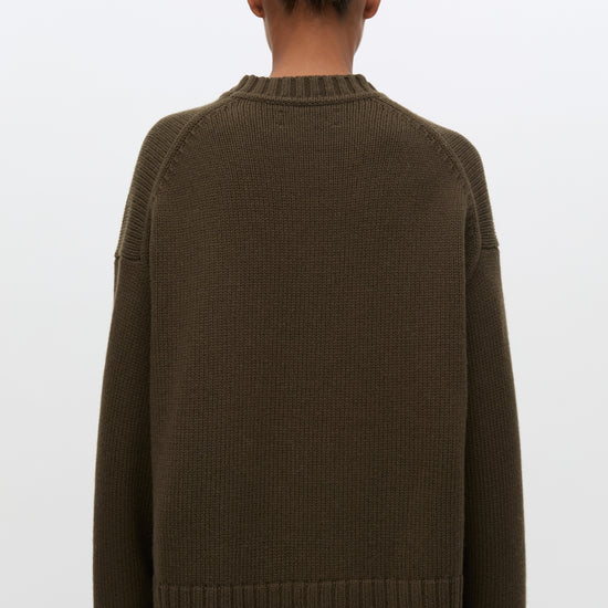 A person shown from the back wearing an oversized ribbed knit sweater in a dark olive brown color, paired with black pants, against a plain light gray studio background.