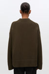 A person shown from the back wearing an oversized ribbed knit sweater in a dark olive brown color, paired with black pants, against a plain light gray studio background.