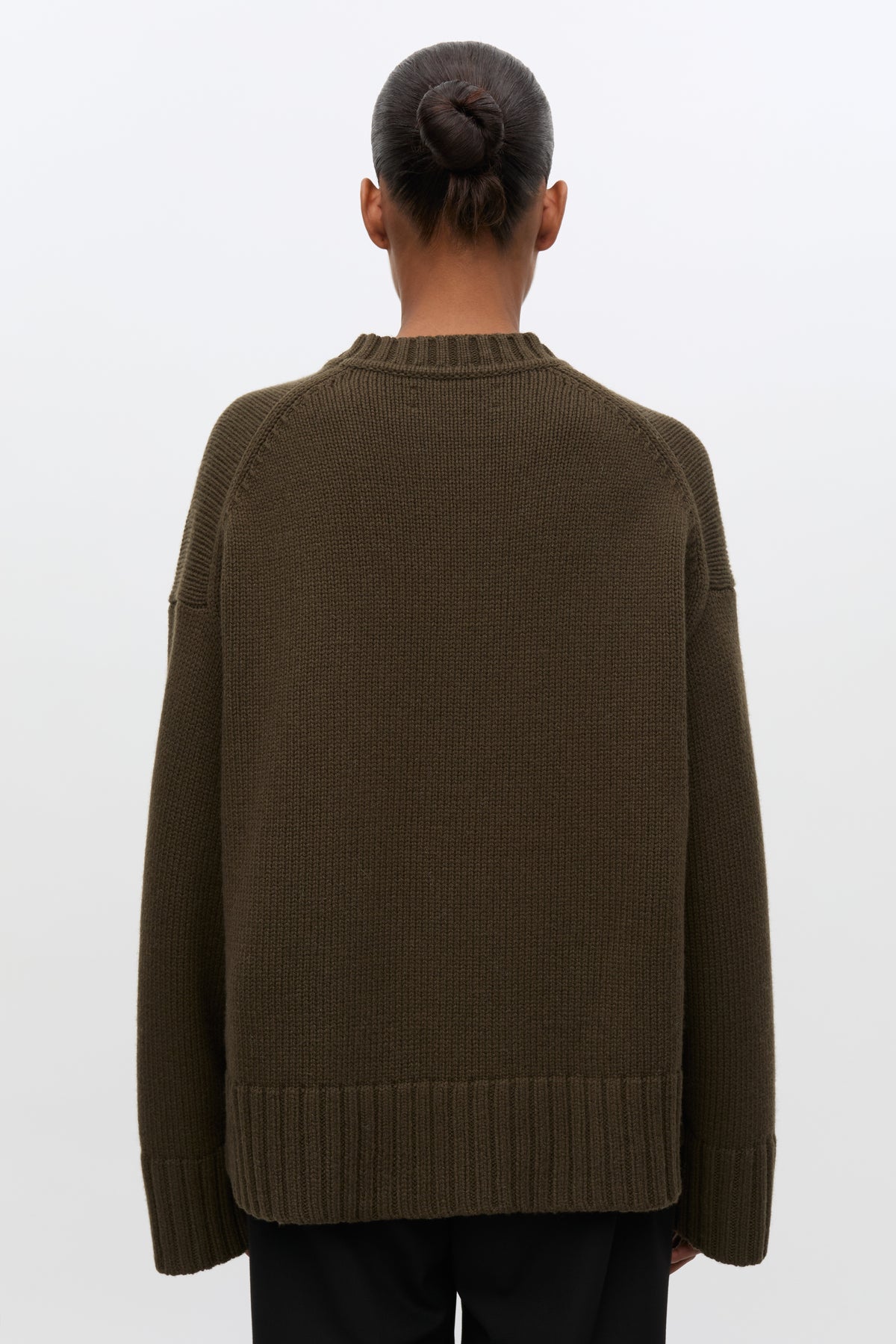 A person shown from the back wearing an oversized ribbed knit sweater in a dark olive brown color, paired with black pants, against a plain light gray studio background.