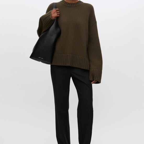 A model stands against a light gray studio background wearing an oversized dark olive-brown knit sweater, black trousers, black shoes, and carrying a large black tote bag.