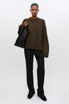 A model stands against a light gray studio background wearing an oversized dark olive-brown knit sweater, black trousers, black shoes, and carrying a large black tote bag.
