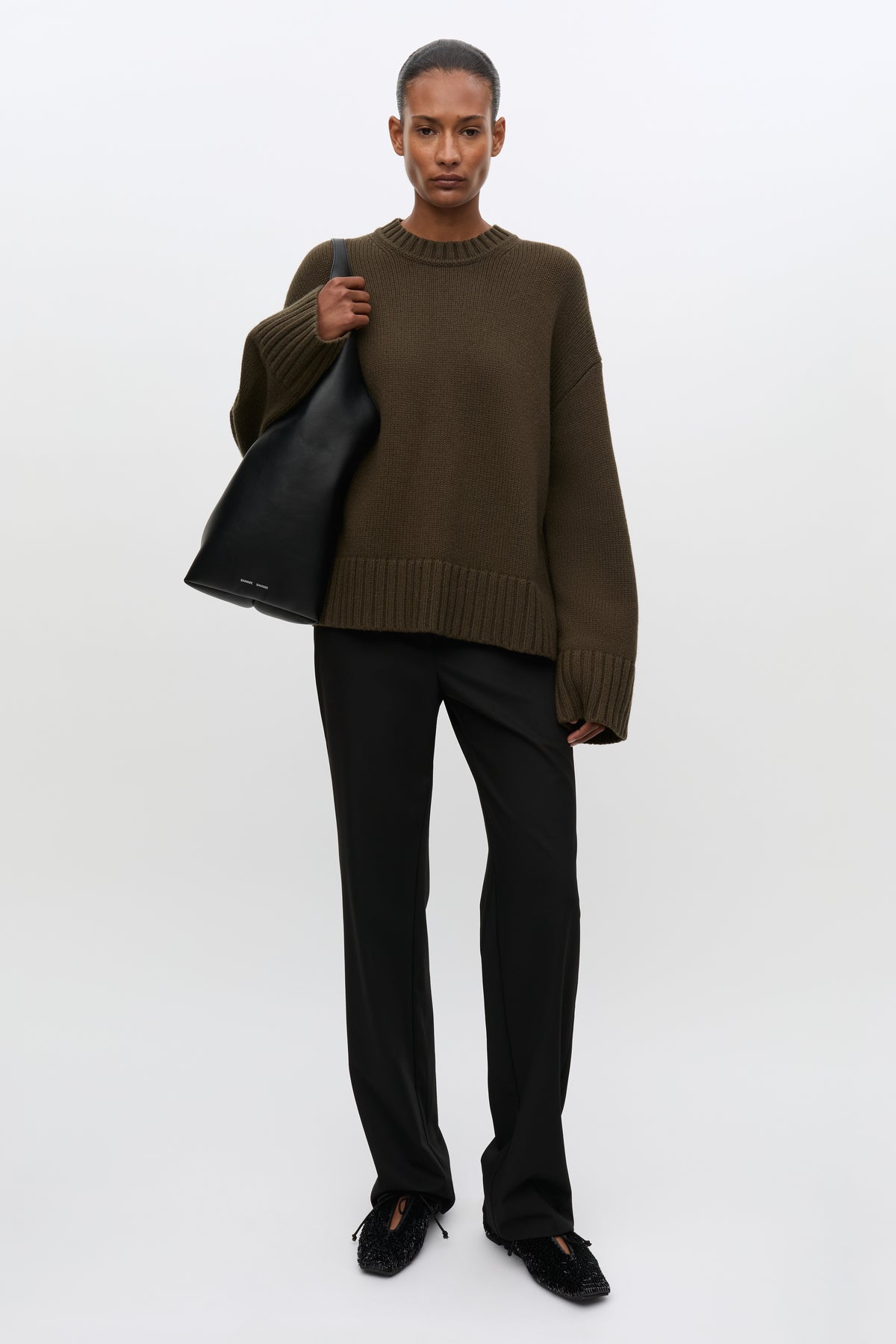 A model stands against a light gray studio background wearing an oversized dark olive-brown knit sweater, black trousers, black shoes, and carrying a large black tote bag.