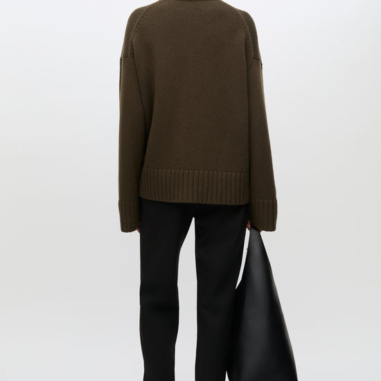 A model shown from the back wearing an oversized dark brown knit sweater, black flared trousers, and holding a large black leather tote bag against a clean white studio background.