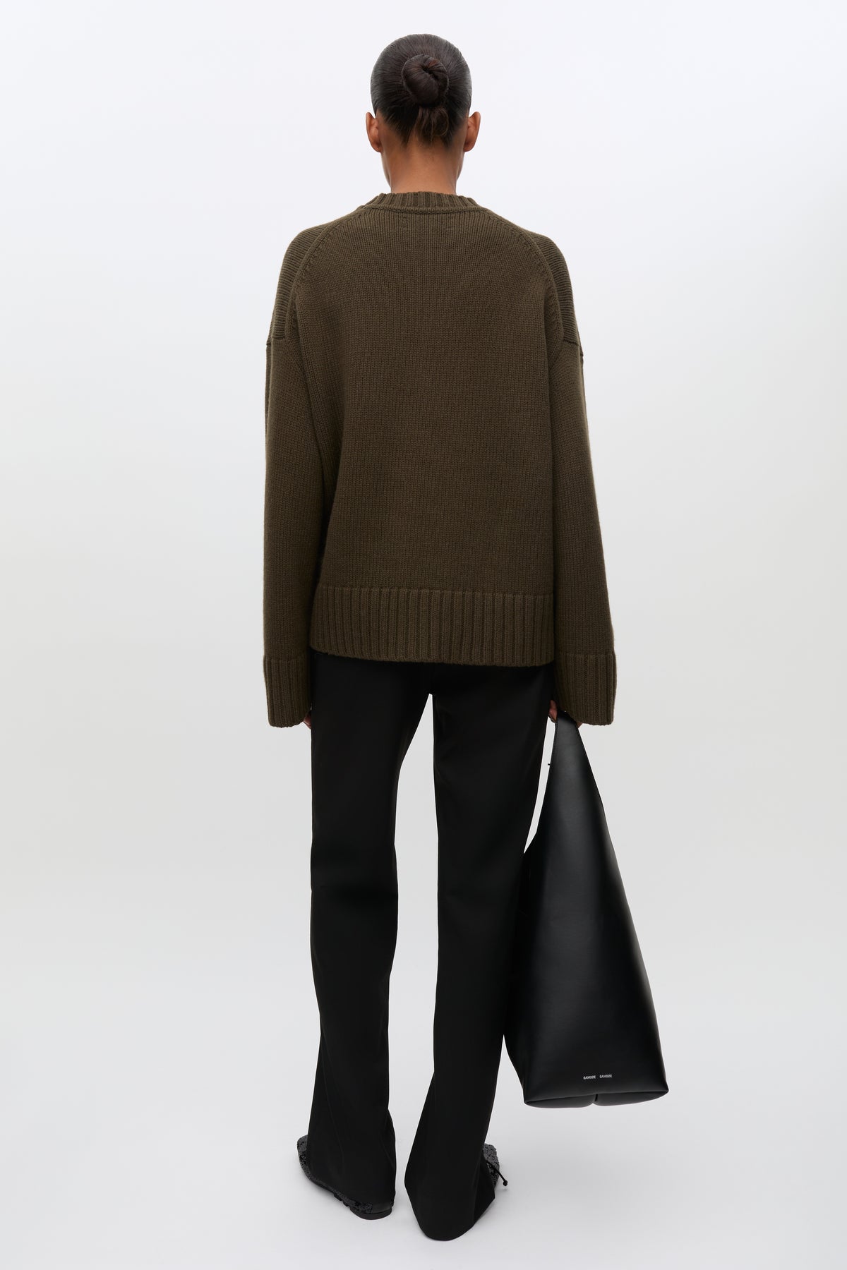 A model shown from the back wearing an oversized dark brown knit sweater, black flared trousers, and holding a large black leather tote bag against a clean white studio background.
