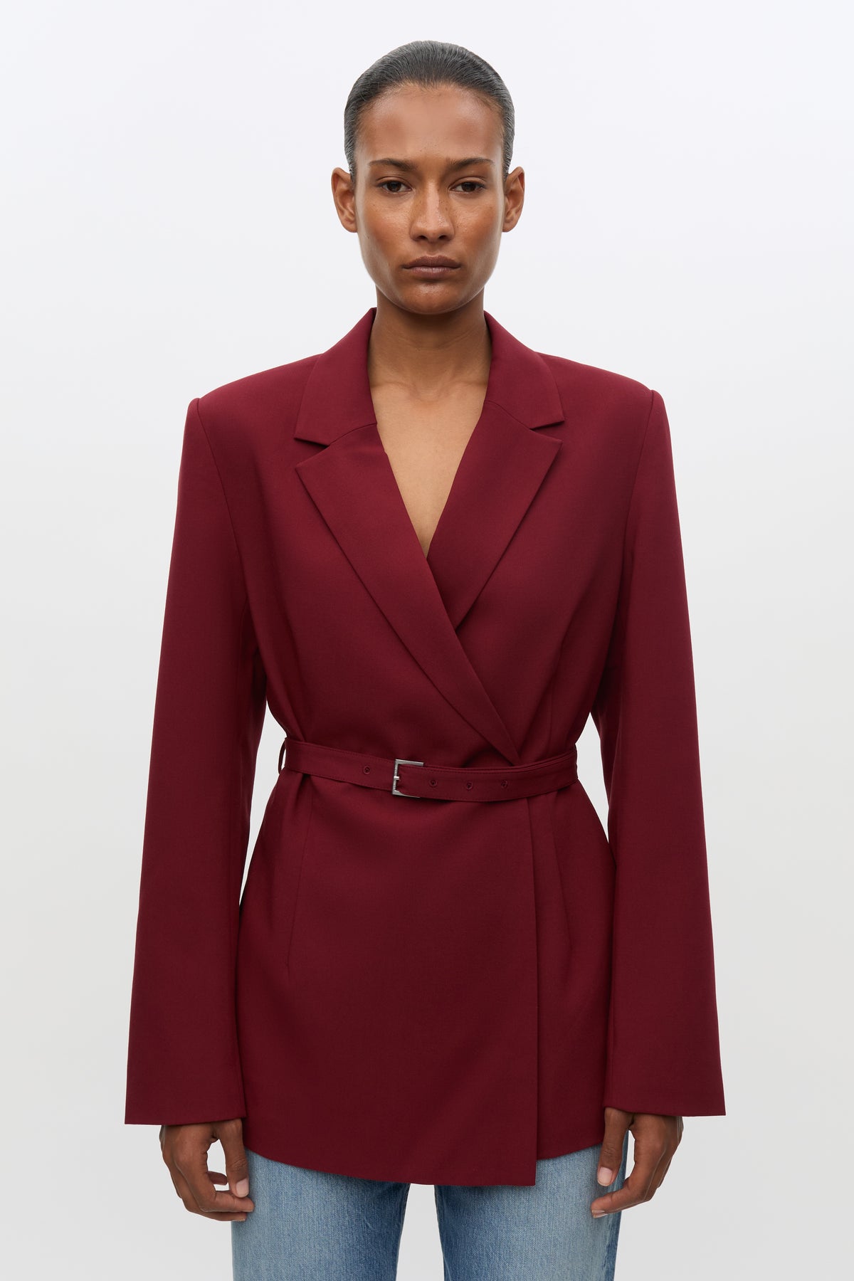 A model wearing a deep burgundy wrap-style blazer with a matching waist belt, paired with light blue jeans, against a plain light gray studio background.