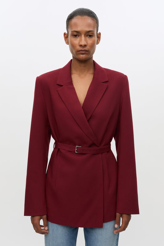 A model wearing a deep burgundy wrap-style blazer with a matching waist belt, paired with light blue jeans, against a plain light gray studio background.