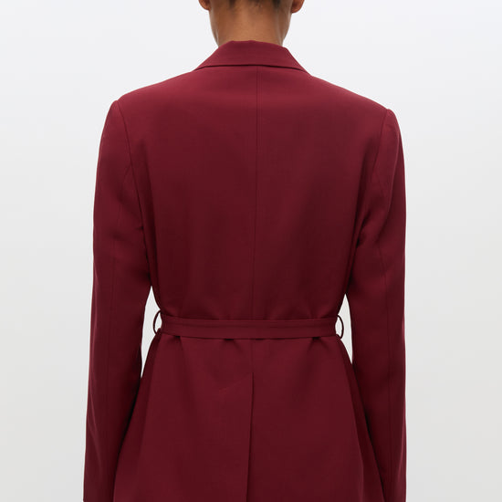 A person shown from the back wearing a tailored blazer in deep burgundy with a matching tie belt at the waist, paired with blue jeans against a plain light gray studio background.