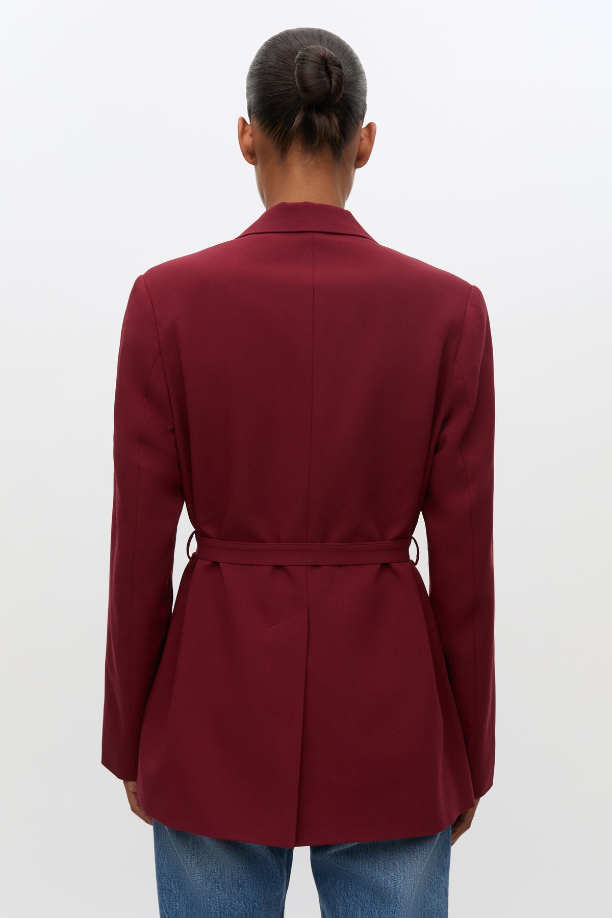 A person shown from the back wearing a tailored blazer in deep burgundy with a matching tie belt at the waist, paired with blue jeans against a plain light gray studio background.