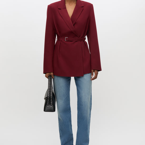 A model stands against a light gray studio background wearing a tailored wrap-style blazer in deep burgundy with a matching waist belt, blue jeans, black shoes, and holding a black handbag.