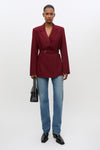 A model stands against a light gray studio background wearing a tailored wrap-style blazer in deep burgundy with a matching waist belt, blue jeans, black shoes, and holding a black handbag.