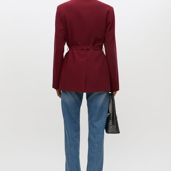 A model shown from the back wearing a structured burgundy belted blazer, blue jeans, and black shoes, holding a black handbag against a plain white studio background.