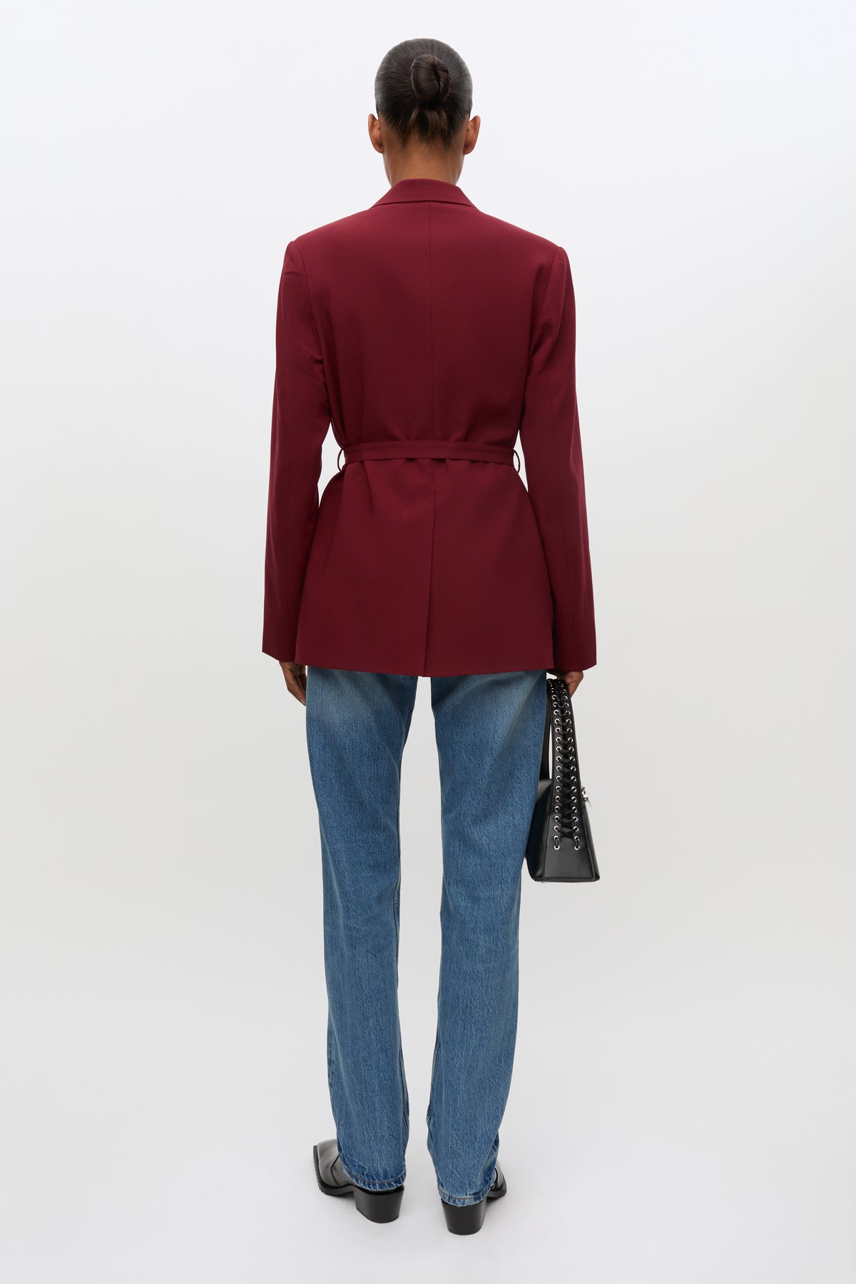 A model shown from the back wearing a structured burgundy belted blazer, blue jeans, and black shoes, holding a black handbag against a plain white studio background.