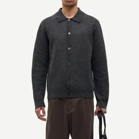 A man modeling a dark gray knit button-up cardigan with a polo collar, paired with brown trousers and holding a black tote bag, against a plain light gray background.