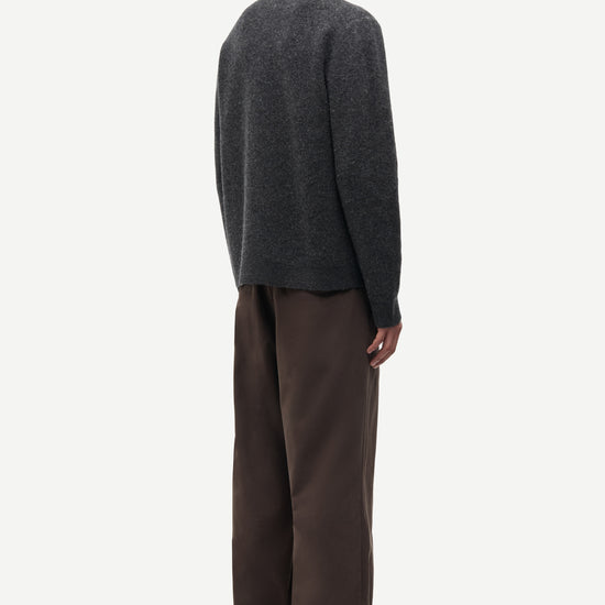 A man shown from the back wearing a dark charcoal gray knit sweater and brown wide-leg trousers, standing against a plain light gray studio background.
