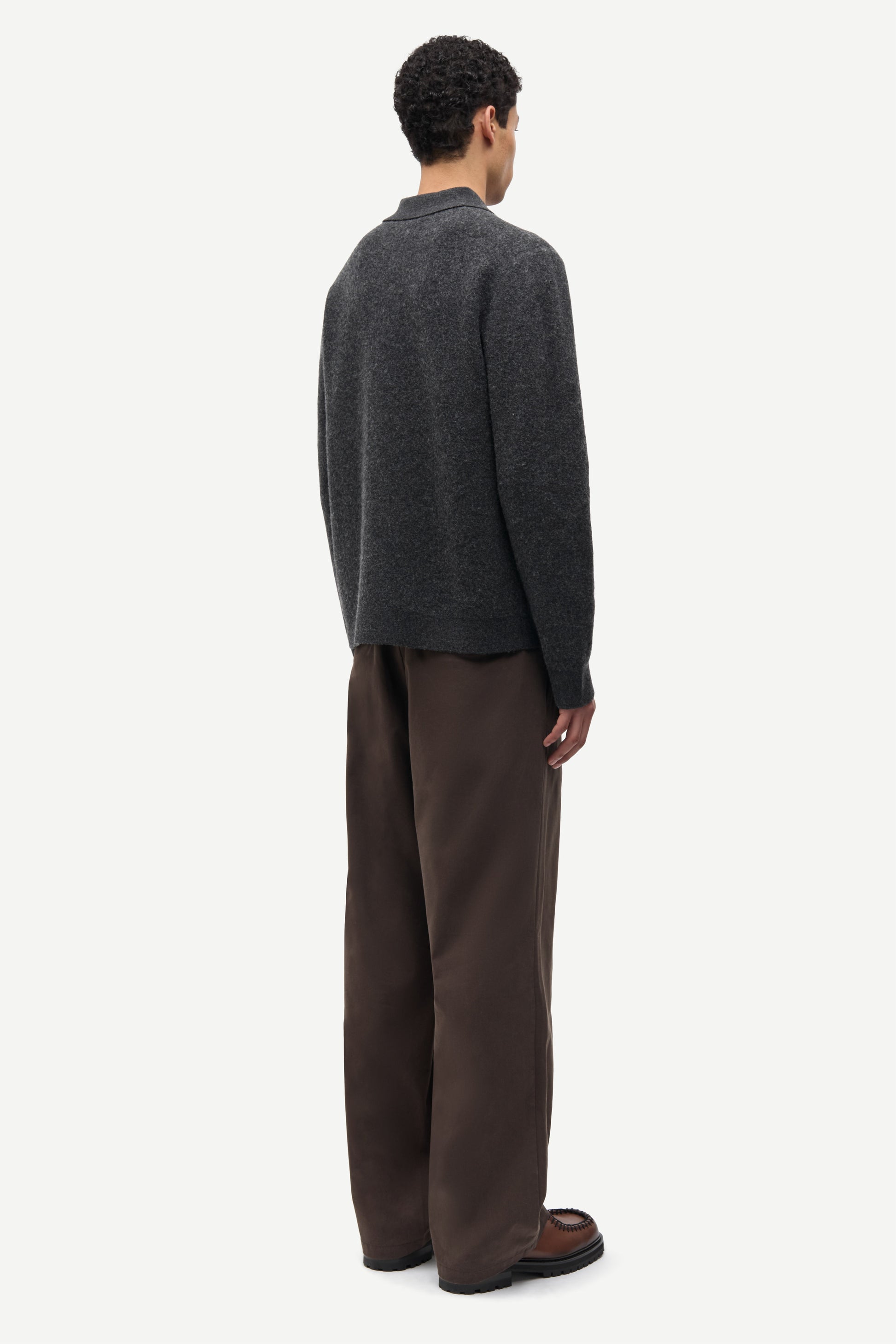 A man shown from the back wearing a dark charcoal gray knit sweater and brown wide-leg trousers, standing against a plain light gray studio background.
