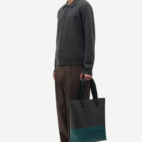 A man stands in profile wearing a dark gray knit polo sweater, brown trousers, and brown shoes, holding a dark tote bag with a teal lower panel. The background is a clean, light off-white studio backdrop.