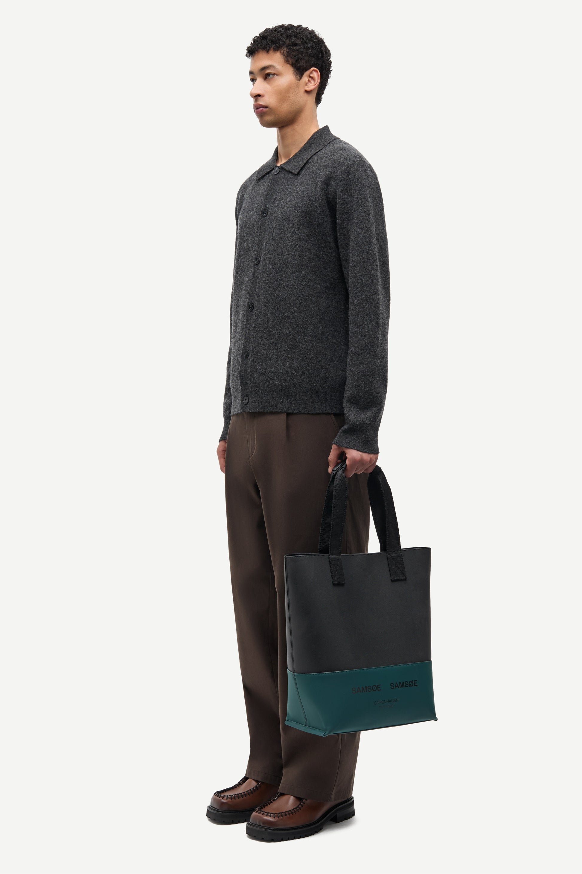 A man stands in profile wearing a dark gray knit polo sweater, brown trousers, and brown shoes, holding a dark tote bag with a teal lower panel. The background is a clean, light off-white studio backdrop.