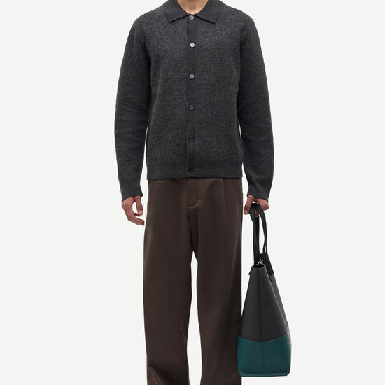 A man stands against a light gray studio background wearing a dark charcoal knit button-front cardigan, brown trousers, and brown loafers, while holding a dark tote bag with teal color blocking.