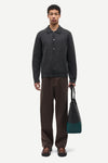 A man stands against a light gray studio background wearing a dark charcoal knit button-front cardigan, brown trousers, and brown loafers, while holding a dark tote bag with teal color blocking.