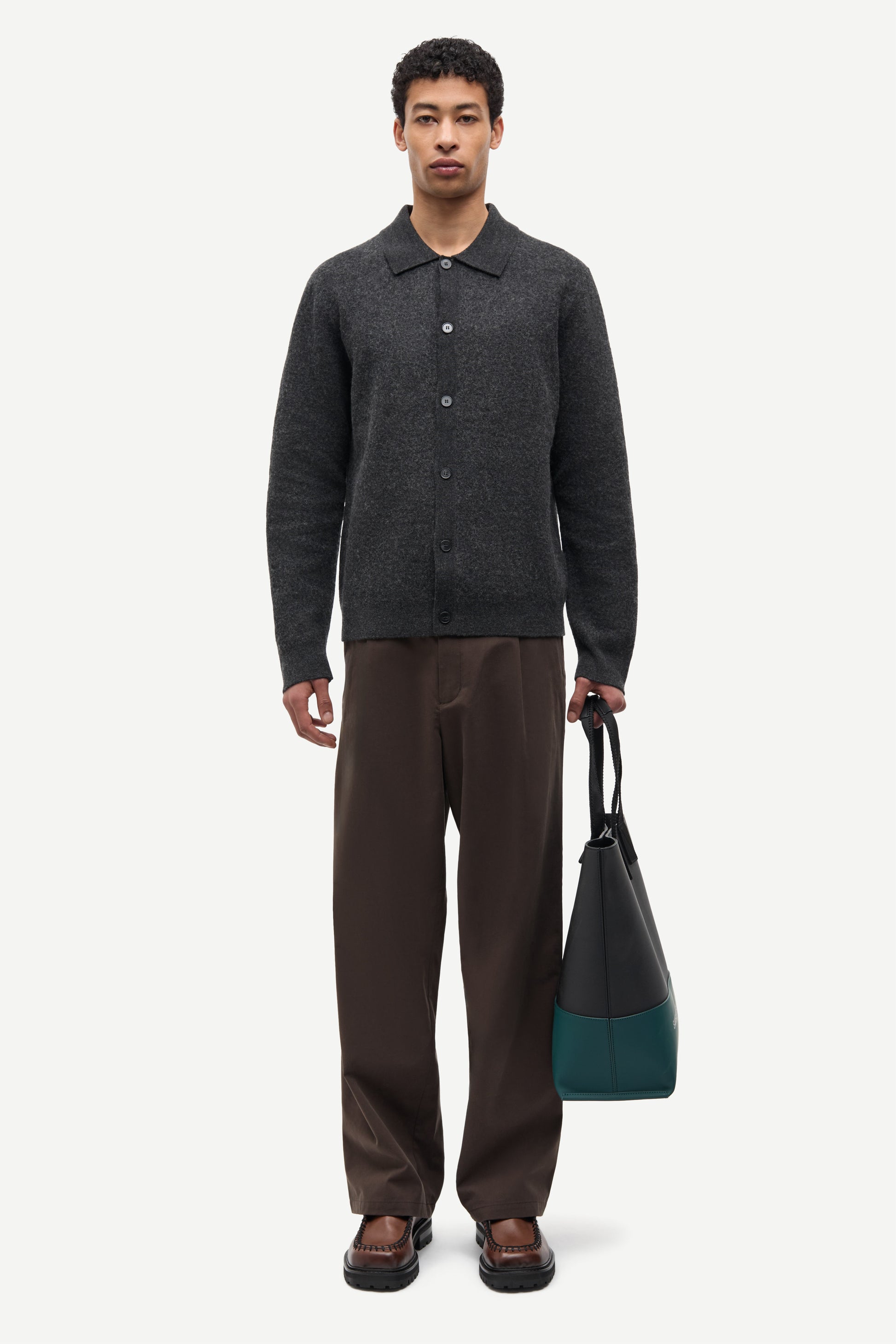 A man stands against a light gray studio background wearing a dark charcoal knit button-front cardigan, brown trousers, and brown loafers, while holding a dark tote bag with teal color blocking.