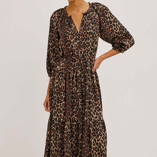 A woman modeling a leopard-print midi dress with puff sleeves and a tiered skirt, photographed against a plain light gray studio background.