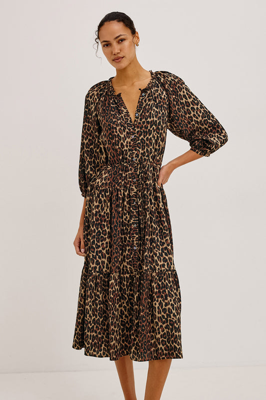 A woman modeling a leopard-print midi dress with puff sleeves and a tiered skirt, photographed against a plain light gray studio background.