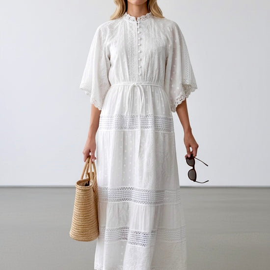 Woman wearing a white dress holding a straw bag and sunglasses against a plain background