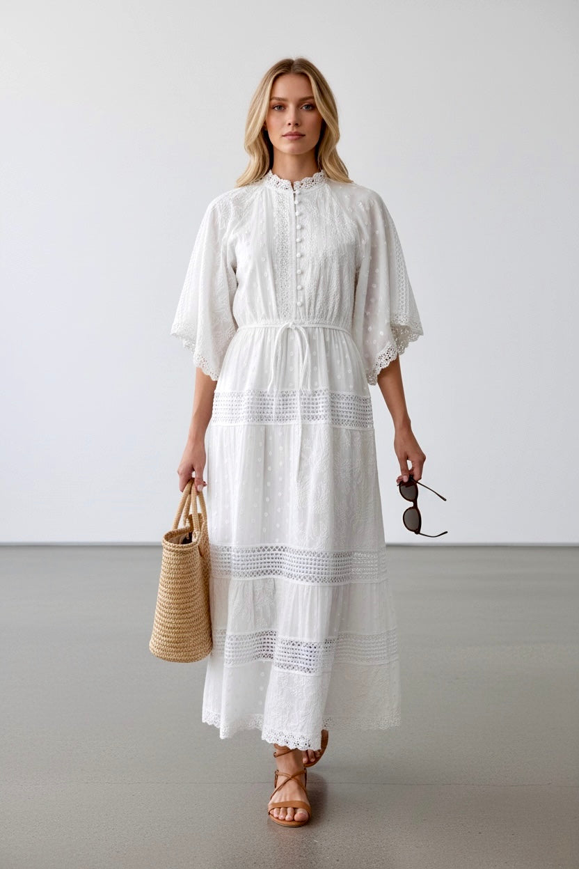 Woman wearing a white dress holding a straw bag and sunglasses against a plain background