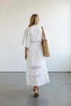 Woman in a white dress holding a woven bag against a plain background
