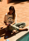 Woman sitting by a pool wearing a 'La Pasta' shirt, holding a phone.